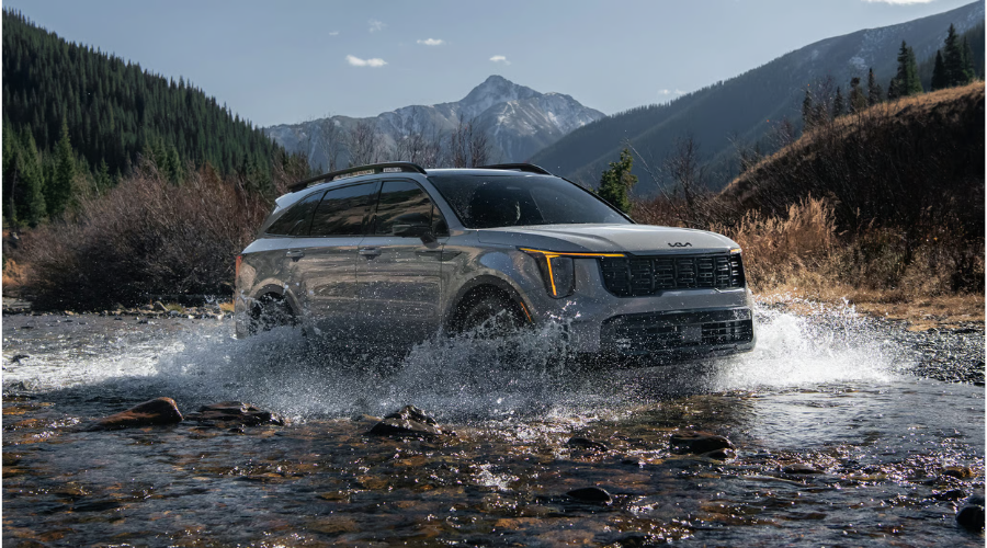 A silver 2026 Kia Sorento splashing through a shallow, rocky riverbed with water spraying, demonstrating its rugged capability and driving dynamics against a backdrop of pine trees and mountains in Waukegan