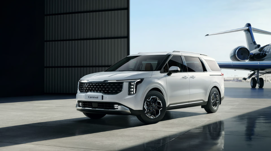 White 2026 Kia Carnival Multi-Purpose Vehicle featuring modern front grille design and alloy wheels outside an aircraft hangar, in stock at Classic Kia serving Waukegan and Lake County drivers