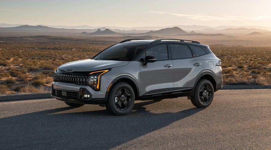 Front three-quarter view of a gray 2026 Kia Sportage crossover on an open road, highlighting its amber LED daytime running lights, black alloy wheels, roof rails, and front radar sensor housing available at Classic Kia near Waukegan, IL.
