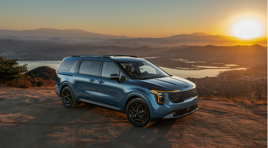 Front three-quarter view of a metallic blue 2027 Kia Carnival multi-purpose vehicle parked at sunset on a scenic overlook, highlighting its SUV-inspired front grille and LED daytime running lights available at Classic Kia in Waukegan, IL.